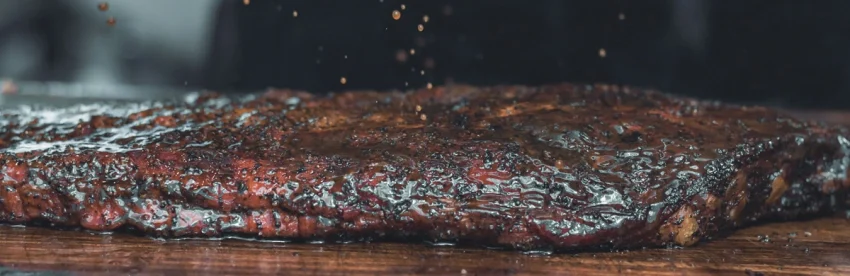 Close-up of a coffee-rubbed brisket with a dark glossy bark crust resting on a wooden cutting board, with spices falling through the air