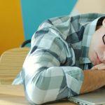 Man in a checked shirt and glasses asleep with his head on his arms at a desk, a steaming turquoise mug of coffee beside his laptop