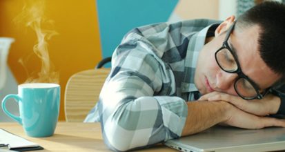Man in a checked shirt and glasses asleep with his head on his arms at a desk, a steaming turquoise mug of coffee beside his laptop