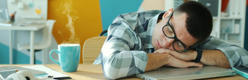 Man in a checked shirt and glasses asleep with his head on his arms at a desk, a steaming turquoise mug of coffee beside his laptop