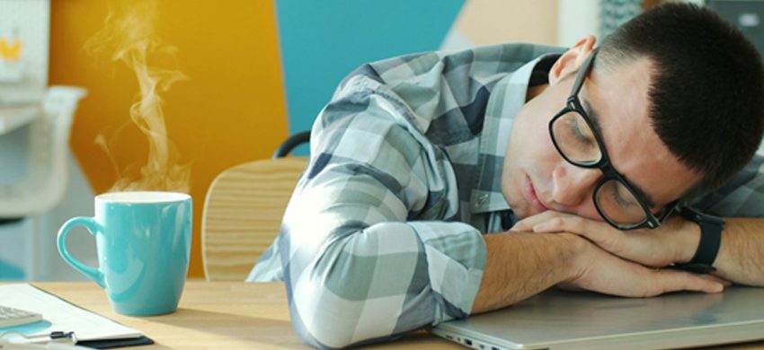 Man in a checked shirt and glasses asleep with his head on his arms at a desk, a steaming turquoise mug of coffee beside his laptop