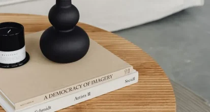 A stack of oversized art and photography coffee table books on a round wooden coffee table, topped with a black candle and vase