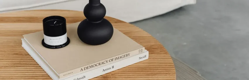 A stack of oversized art and photography coffee table books on a round wooden coffee table, topped with a black candle and vase