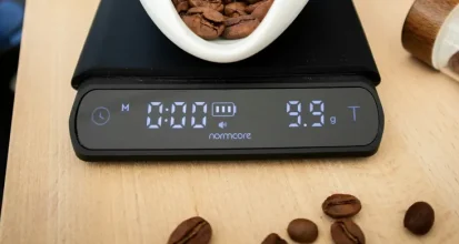 A ceramic dish of coffee beans sitting on a digital kitchen scale reading 9.9 grams, with a coffee grinder and glass canisters nearby