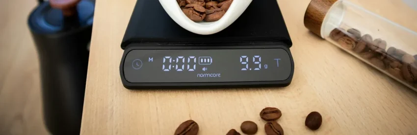 A ceramic dish of coffee beans sitting on a digital kitchen scale reading 9.9 grams, with a coffee grinder and glass canisters nearby