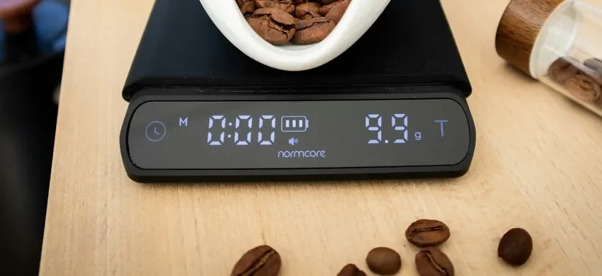 A ceramic dish of coffee beans sitting on a digital kitchen scale reading 9.9 grams, with a coffee grinder and glass canisters nearby
