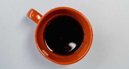 Overhead view of a burnt orange ceramic mug of black coffee gone cold and still, sitting on a plain pale grey surface
