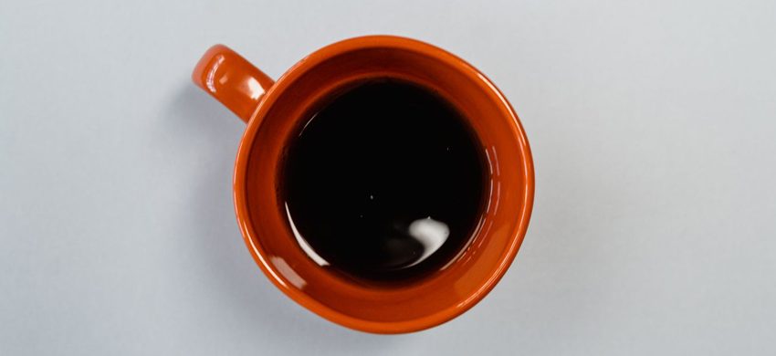 Overhead view of a burnt orange ceramic mug of black coffee gone cold and still, sitting on a plain pale grey surface