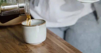 A person pouring freshly brewed pour-over coffee from a glass carafe into a ceramic mug in a home kitchen