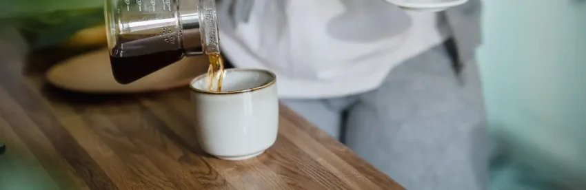 A person pouring freshly brewed pour-over coffee from a glass carafe into a ceramic mug in a home kitchen
