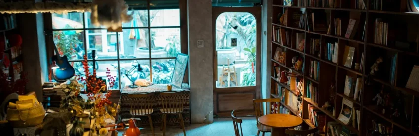 The warm, characterful interior of an independent coffee shop, with floor-to-ceiling bookshelves, mismatched furniture, and daylight streaming through a large window