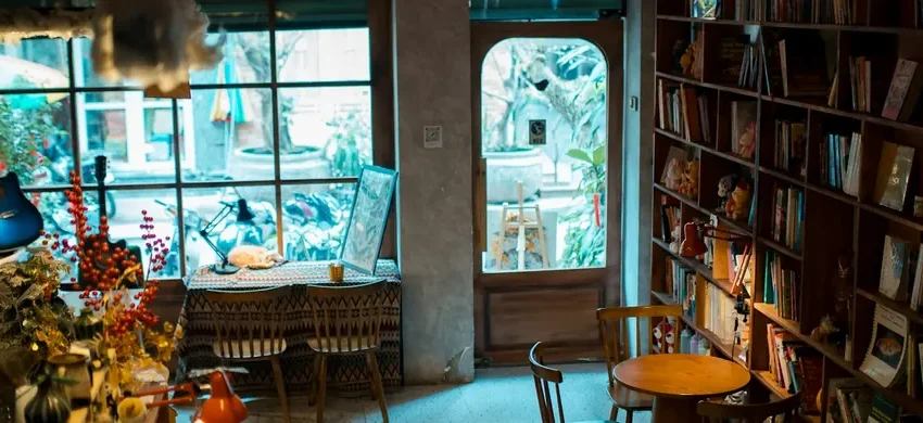 The warm, characterful interior of an independent coffee shop, with floor-to-ceiling bookshelves, mismatched furniture, and daylight streaming through a large window