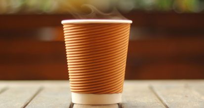 Steam rising from a ribbed brown takeaway coffee cup with a white lid, standing on a weathered wooden table