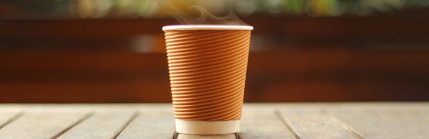 Steam rising from a ribbed brown takeaway coffee cup with a white lid, standing on a weathered wooden table