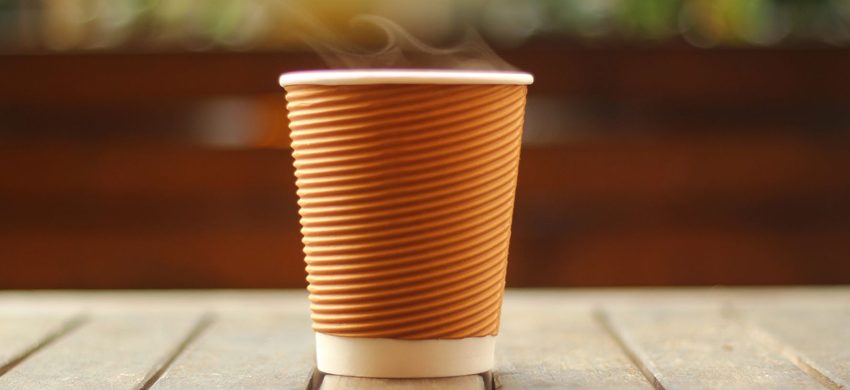 Steam rising from a ribbed brown takeaway coffee cup with a white lid, standing on a weathered wooden table