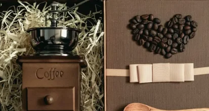 A manual coffee grinder nestled in a gift box with tissue paper, next to a heart shape made of coffee beans, a ribbon, and a wooden spoon