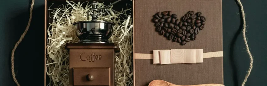 A manual coffee grinder nestled in a gift box with tissue paper, next to a heart shape made of coffee beans, a ribbon, and a wooden spoon