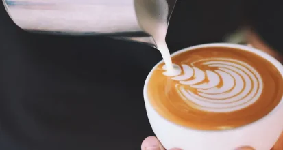 Steamed milk being poured from a metal pitcher into a white cup, creating a rosetta latte art pattern on top of the espresso
