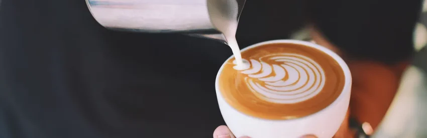 Steamed milk being poured from a metal pitcher into a white cup, creating a rosetta latte art pattern on top of the espresso