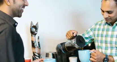 Two coworkers at an office break room coffee station, one pouring fresh coffee from a carafe into a mug for the other