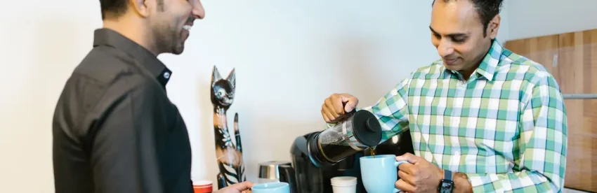 Two coworkers at an office break room coffee station, one pouring fresh coffee from a carafe into a mug for the other