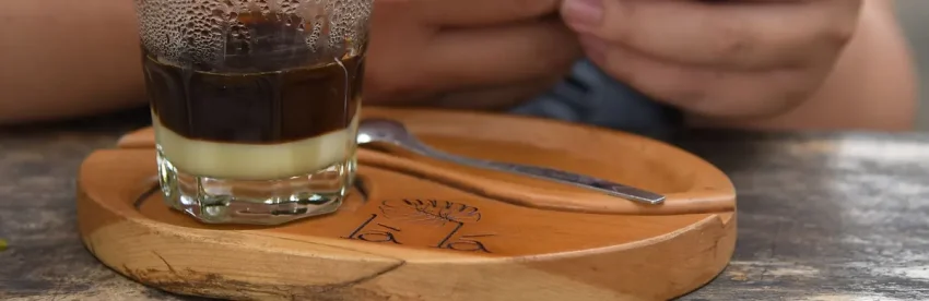 A phin filter sitting on a glass of Vietnamese coffee, with dark coffee visibly layered on top of sweetened condensed milk, on a wooden tray