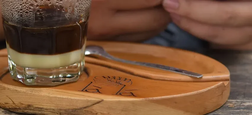 A phin filter sitting on a glass of Vietnamese coffee, with dark coffee visibly layered on top of sweetened condensed milk, on a wooden tray