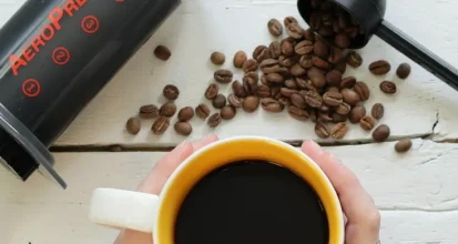 An AeroPress coffee maker lying on a white wooden table beside a scoop of coffee beans, with hands holding a yellow mug of black coffee