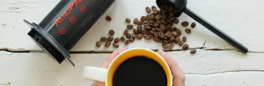 An AeroPress coffee maker lying on a white wooden table beside a scoop of coffee beans, with hands holding a yellow mug of black coffee