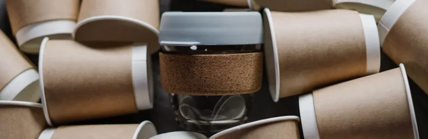 A single reusable coffee cup with a cork sleeve surrounded by dozens of discarded disposable paper coffee cups, illustrating the waste difference