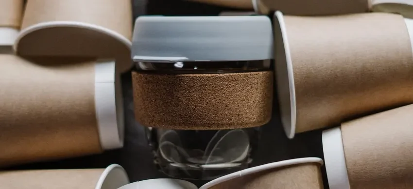 A single reusable coffee cup with a cork sleeve surrounded by dozens of discarded disposable paper coffee cups, illustrating the waste difference