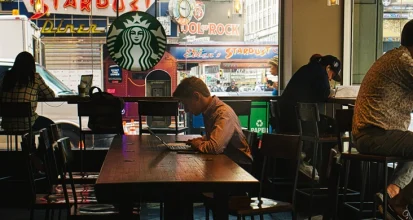 Interior of a busy city Starbucks with several customers working alone on laptops at wooden tables and counter seating, the Starbucks siren logo visible in the window