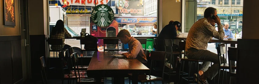 Interior of a busy city Starbucks with several customers working alone on laptops at wooden tables and counter seating, the Starbucks siren logo visible in the window