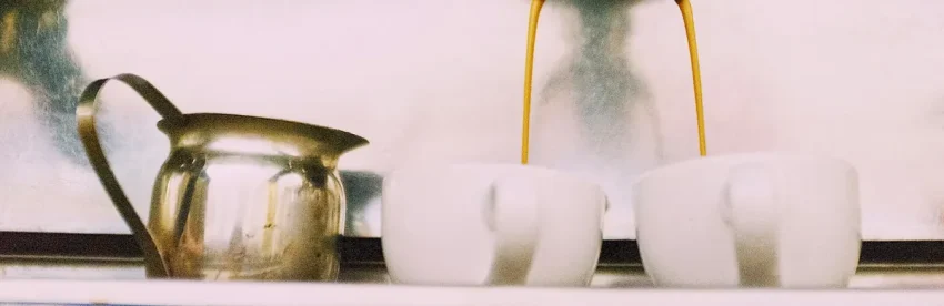 Espresso streaming from the group head of a vintage-style brass espresso machine into two white cups, with a milk pitcher beside them